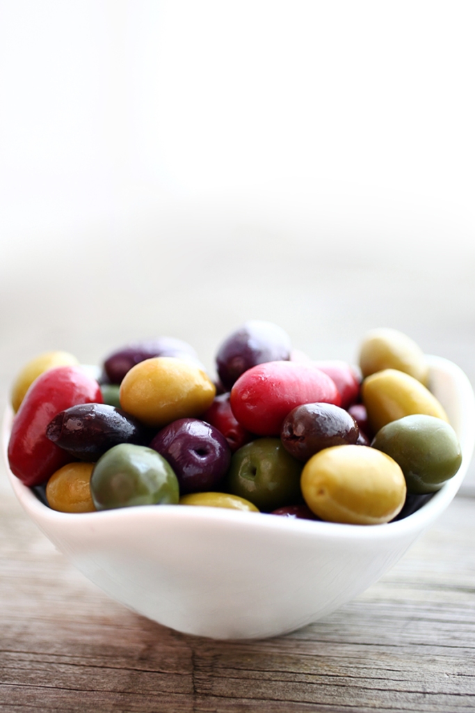 This is an image of a variety of different colored olives in a small white bowl, white window background.